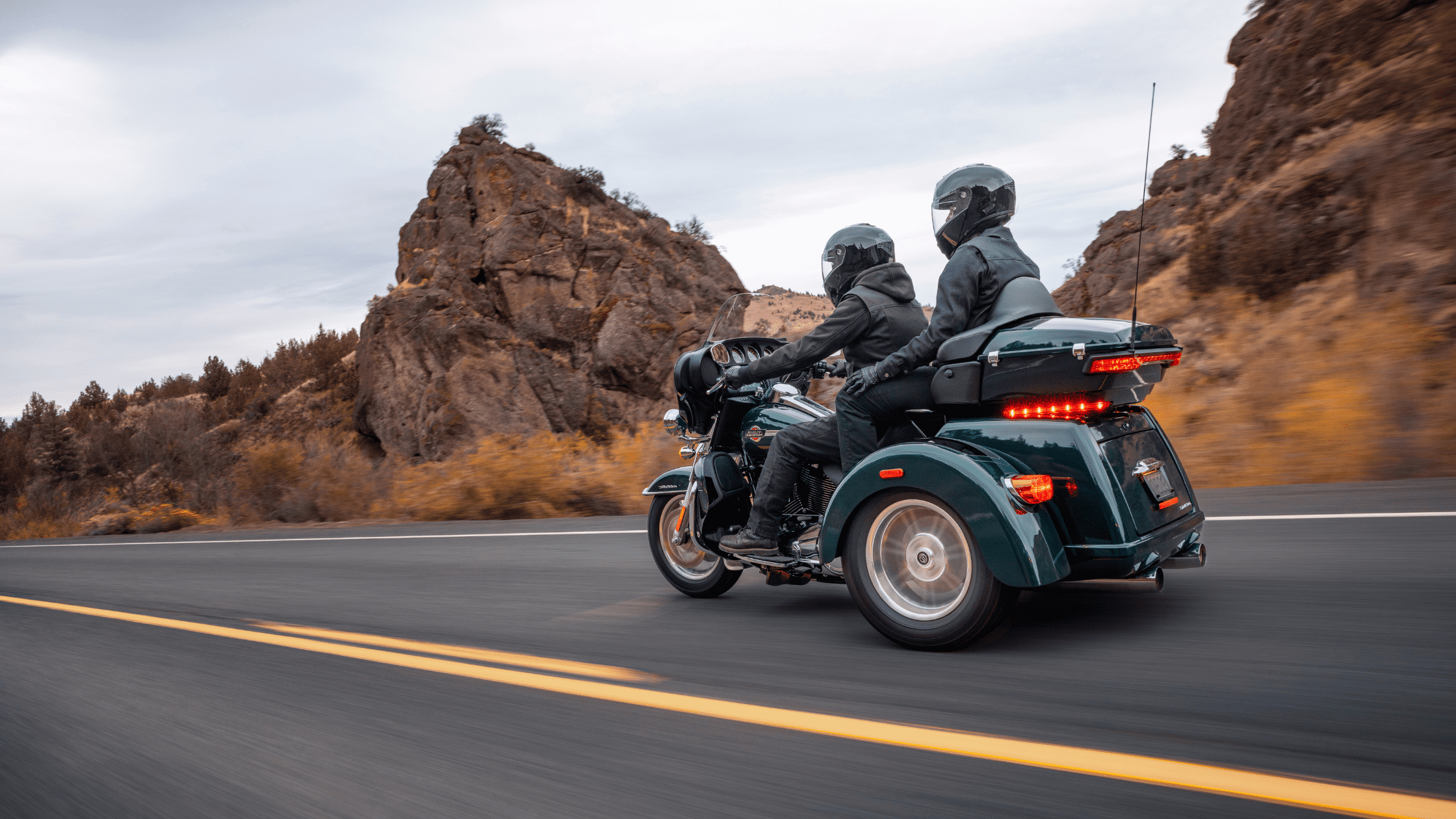 A couple riding a Harley-Davidson Tri Glide Ultra motorcycle on a scenic highway with rocky mountains in the background.