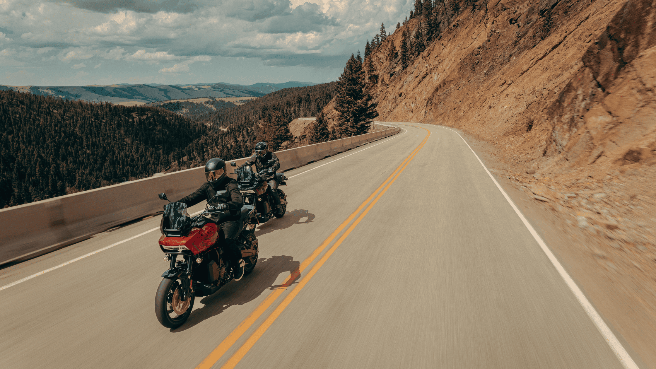 Two adventure touring motorcyclists ride along a winding mountain road, surrounded by rugged cliffs and dense forests. The scenic backdrop emphasizes the thrill of exploration and long-distance touring.