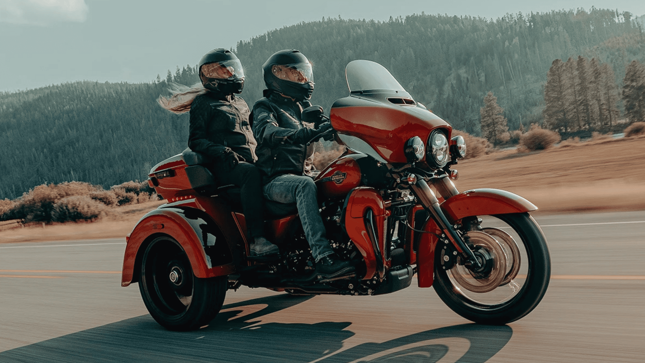 A rider and passenger cruise on a bold red trike motorcycle, set against a backdrop of rolling hills and open countryside. The three-wheeled design ensures stability and comfort for extended road trips.