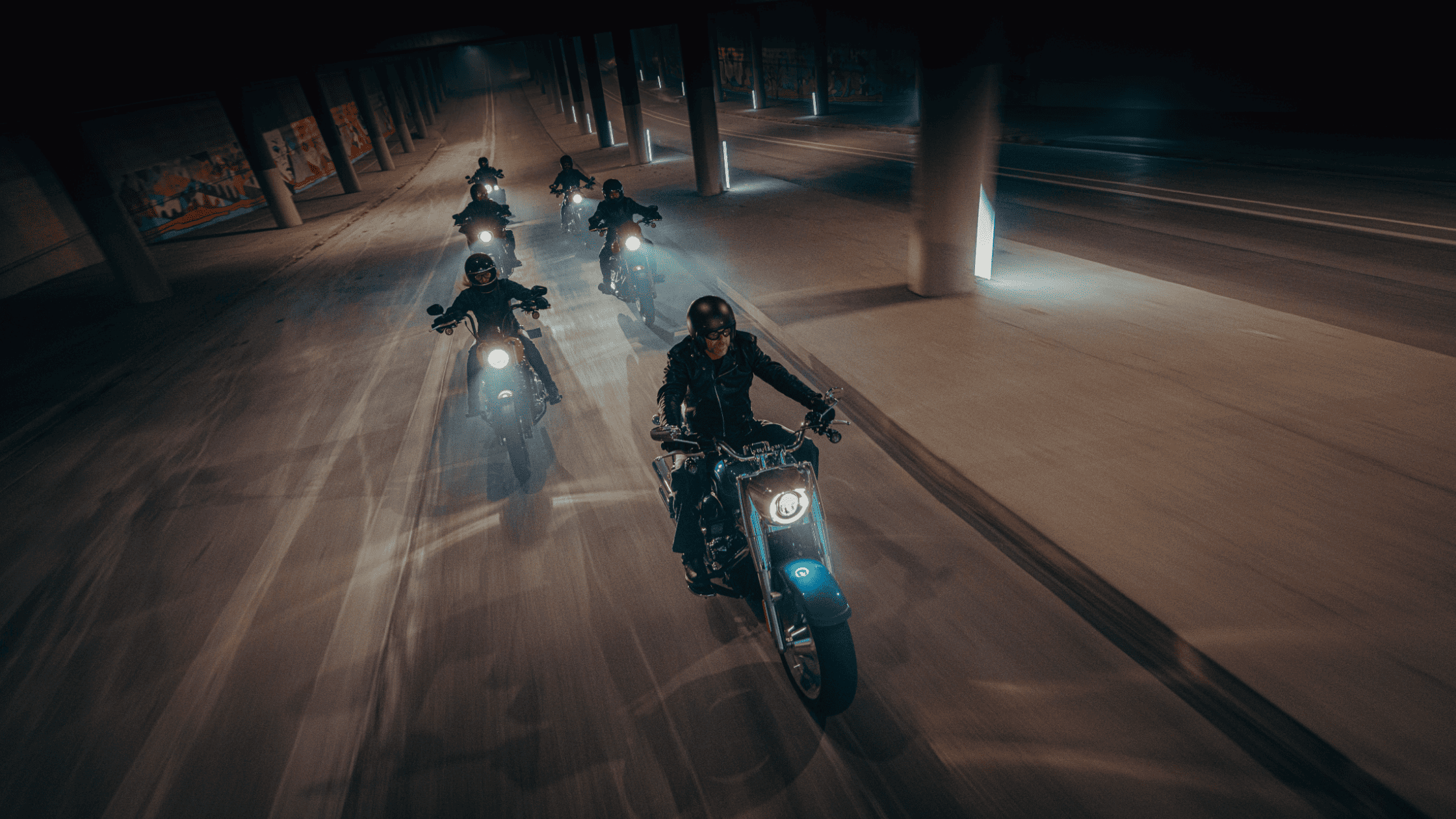A group of riders on cruiser motorcycles powers through a dimly lit urban tunnel, their headlights cutting through the darkness. The scene captures the raw energy and camaraderie of classic motorcycle culture.